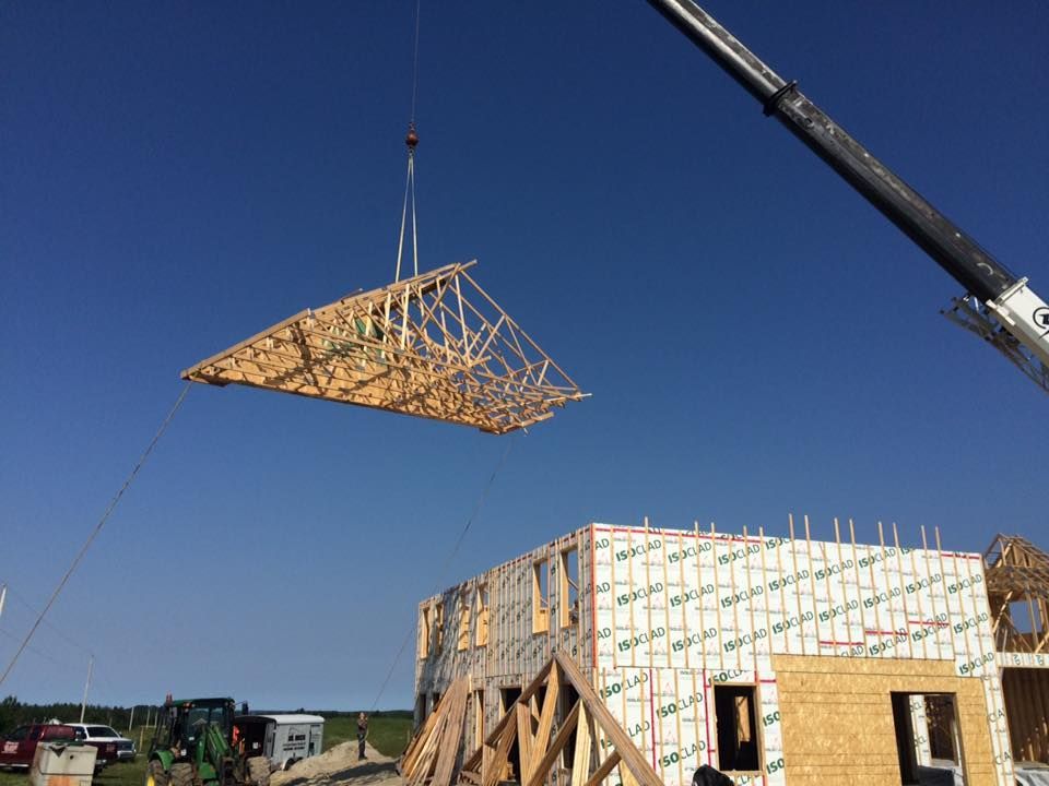 Une grue soulève une ferme de toit en bois au-dessus du chantier d'une maison aux panneaux isolants structurels blancs.