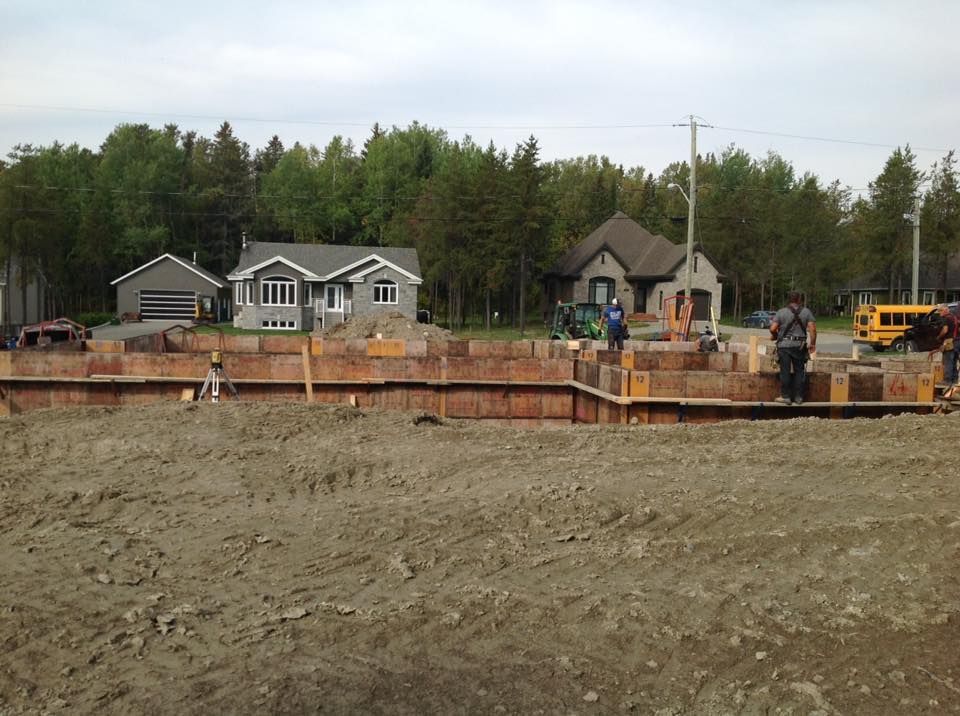 Une équipe de construction installe des coffrages en bois pour les fondations d'une maison sur un terrain vague, avec des maisons voisines à l'arrière.