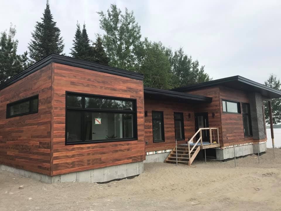 Une maison moderne de plain-pied, revêtue de bois, avec un toit plat, de grandes fenêtres à cadre noir et des fondations en béton.