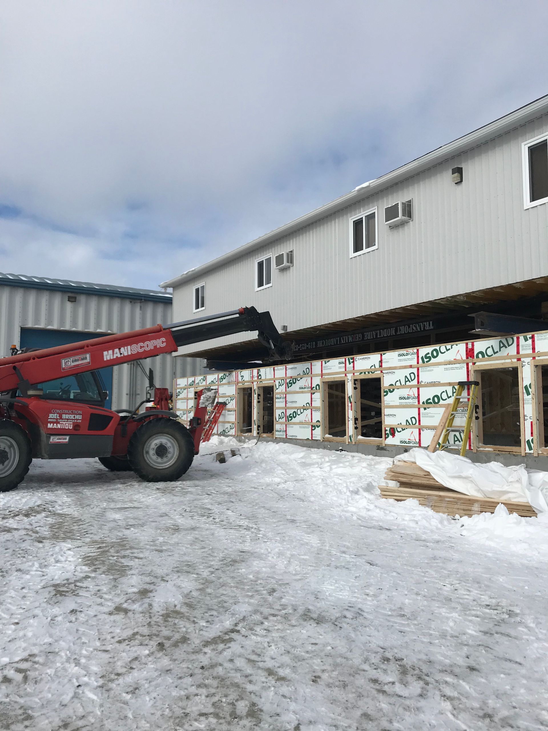 Un chariot télescopique Manitou rouge soulève une poutre d'acier le long du mur extérieur d'un bâtiment en construction sous la neige.