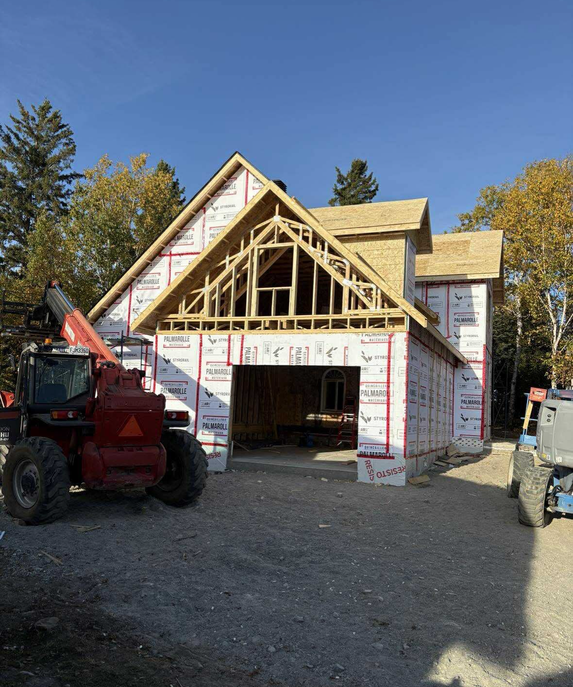 Une maison en construction, avec une charpente en bois apparente et un revêtement blanc résistant aux intempéries, vue depuis une allée de gravier.