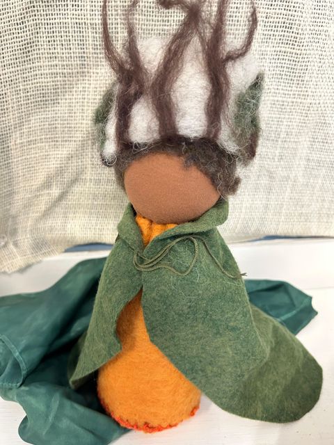 Small handmade doll in a green coat and orange outfit against a patterned fabric background