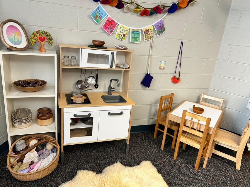 Small play kitchen corner with shelves, wooden tables, chairs, baskets, and colorful wall decorations