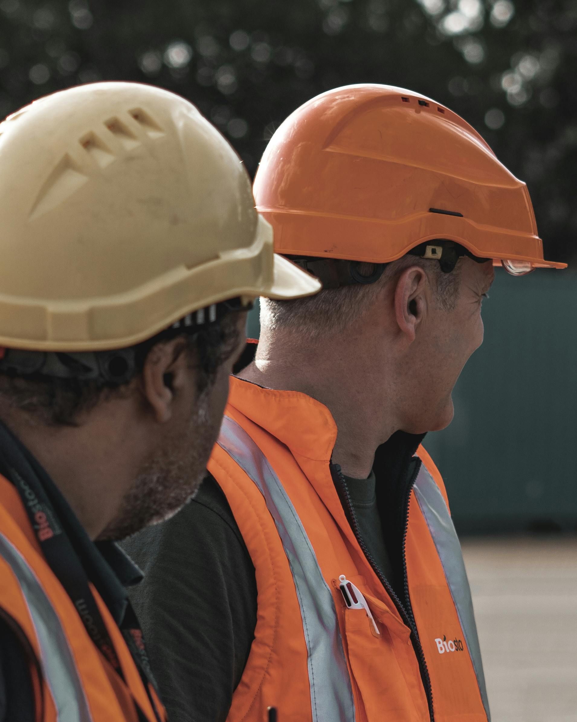 Two construction workers wearing orange safety vests and hard hats (one yellow, one orange) look off to the side.