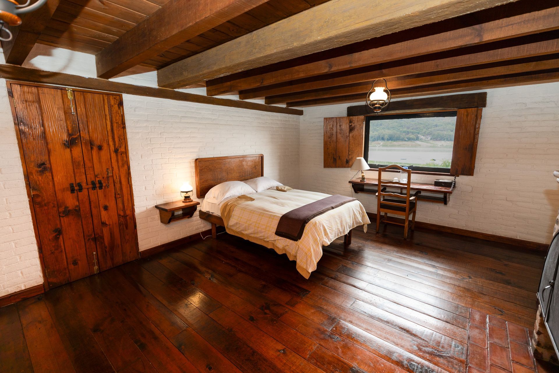 Habitación rústica con vigas de madera, cama, armario y una gran ventana con vistas a la vegetación.