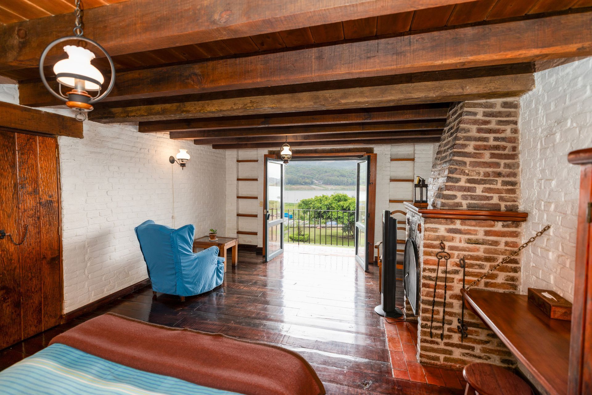 Habitación rústica con vigas vistas, chimenea de ladrillo, silla azul y puertas abiertas con vistas al jardín.