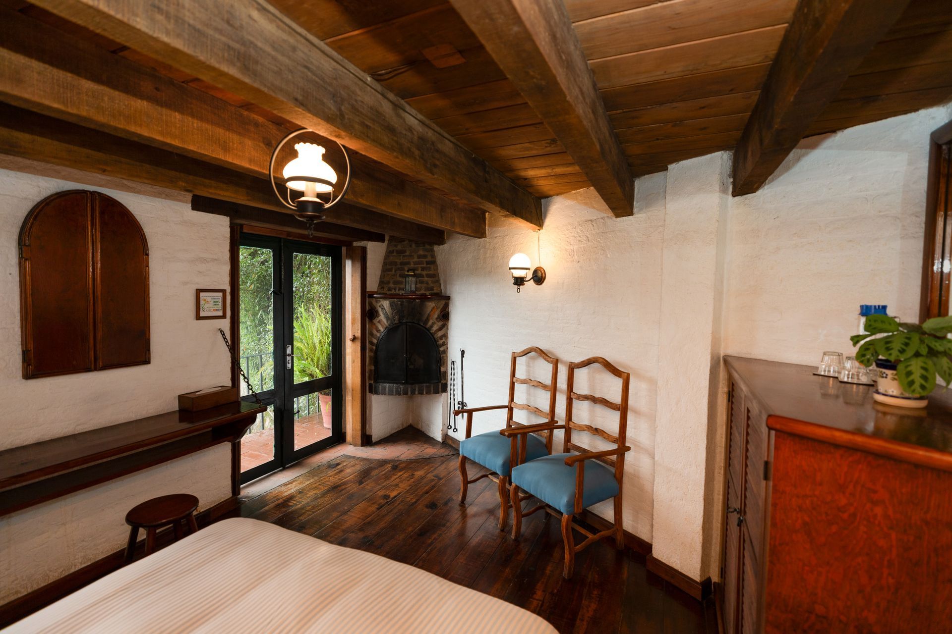 Acogedora habitación rústica con vigas de madera, cama, sillas, chimenea y puertas de cristal que dan a un balcón.