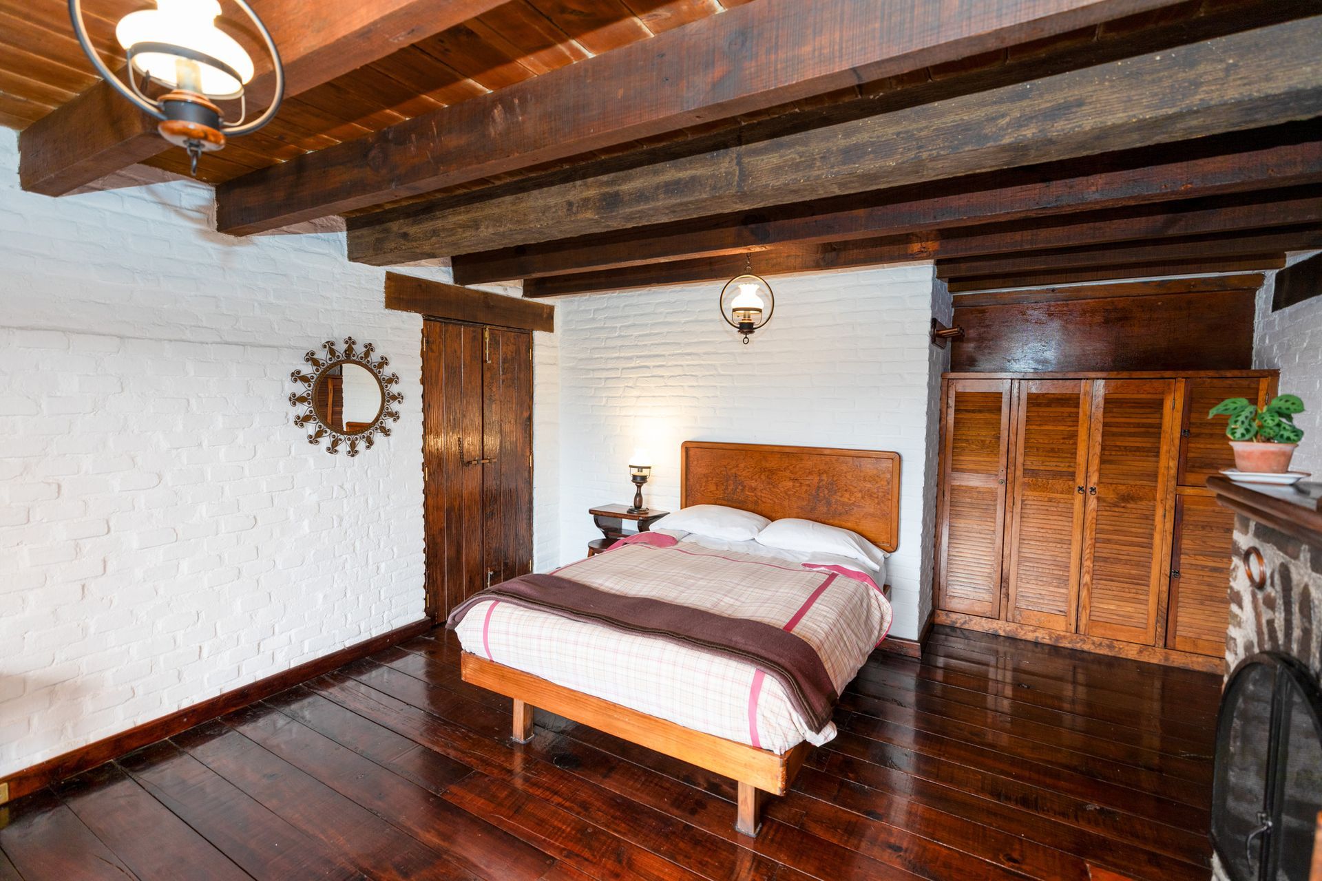 Dormitorio rústico con vigas de madera, paredes de ladrillo blanco, suelo de madera oscura y una cama con ropa de cama rosa.