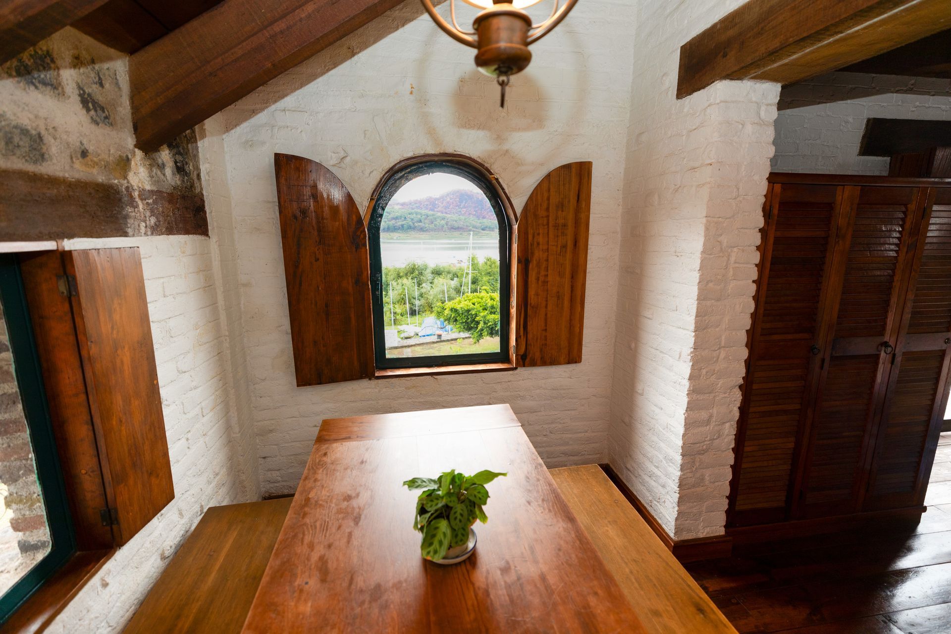 Comedor rústico con mesa de madera, ventana arqueada y luz solar sobre paredes blancas.