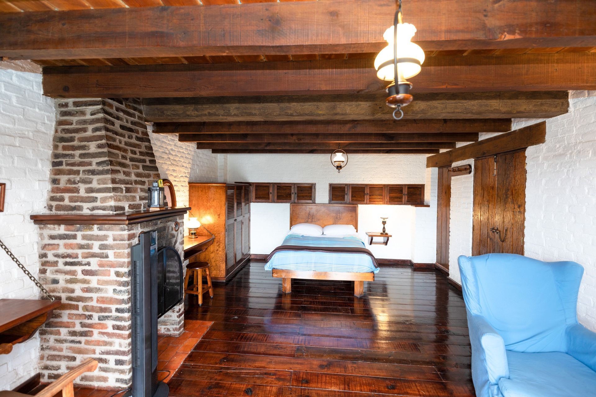 Dormitorio rústico con vigas vistas, chimenea de ladrillo, cama de madera y sillón azul.