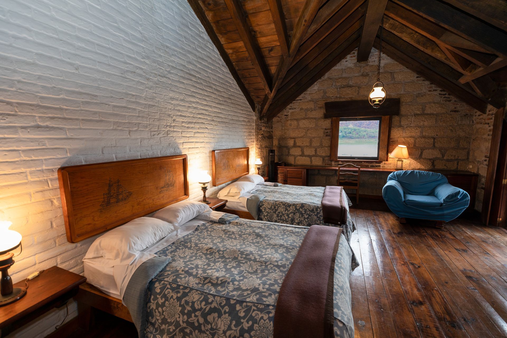 Acogedora habitación abuhardillada con dos camas, vigas de madera, pared de piedra y una silla azul junto a la ventana.