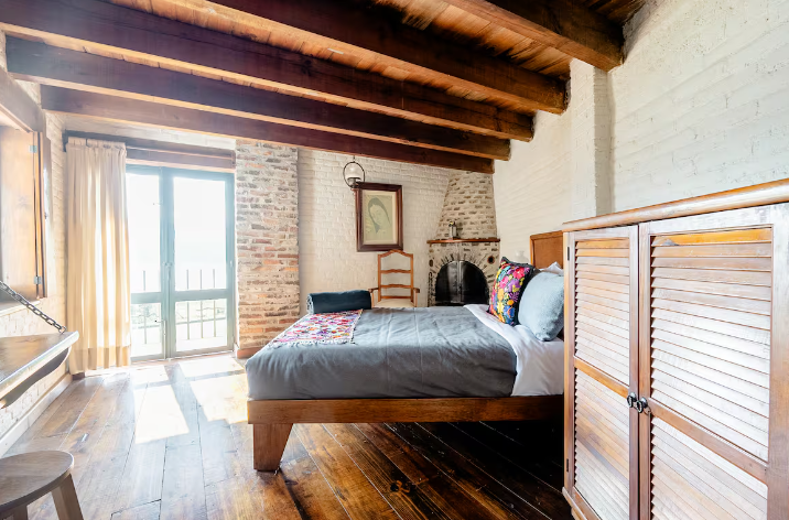 Habitación rústica y luminosa con vigas de madera, cama y puertas que dan al balcón, bañada por la cálida luz del sol.