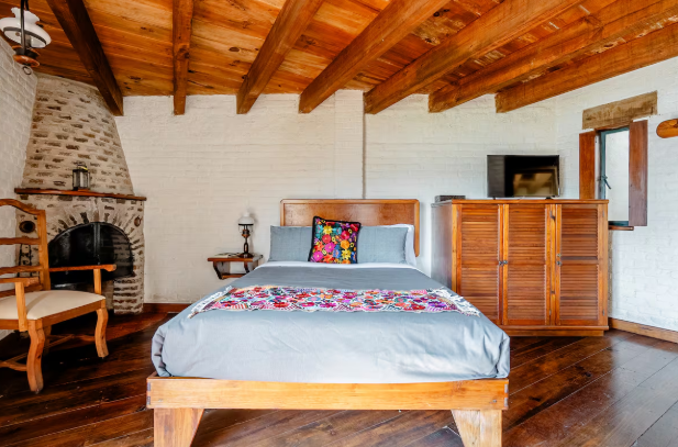 Dormitorio rústico con techo de madera, chimenea de ladrillo, cama y cómoda con televisor.