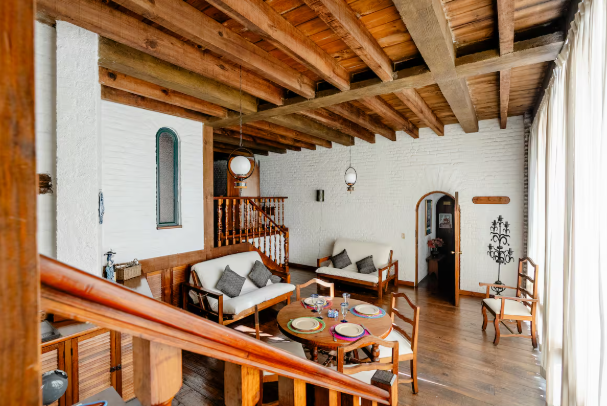 Sala de estar con vigas de madera, sofás, mesa de centro y escalera en un interior rústico y luminoso.