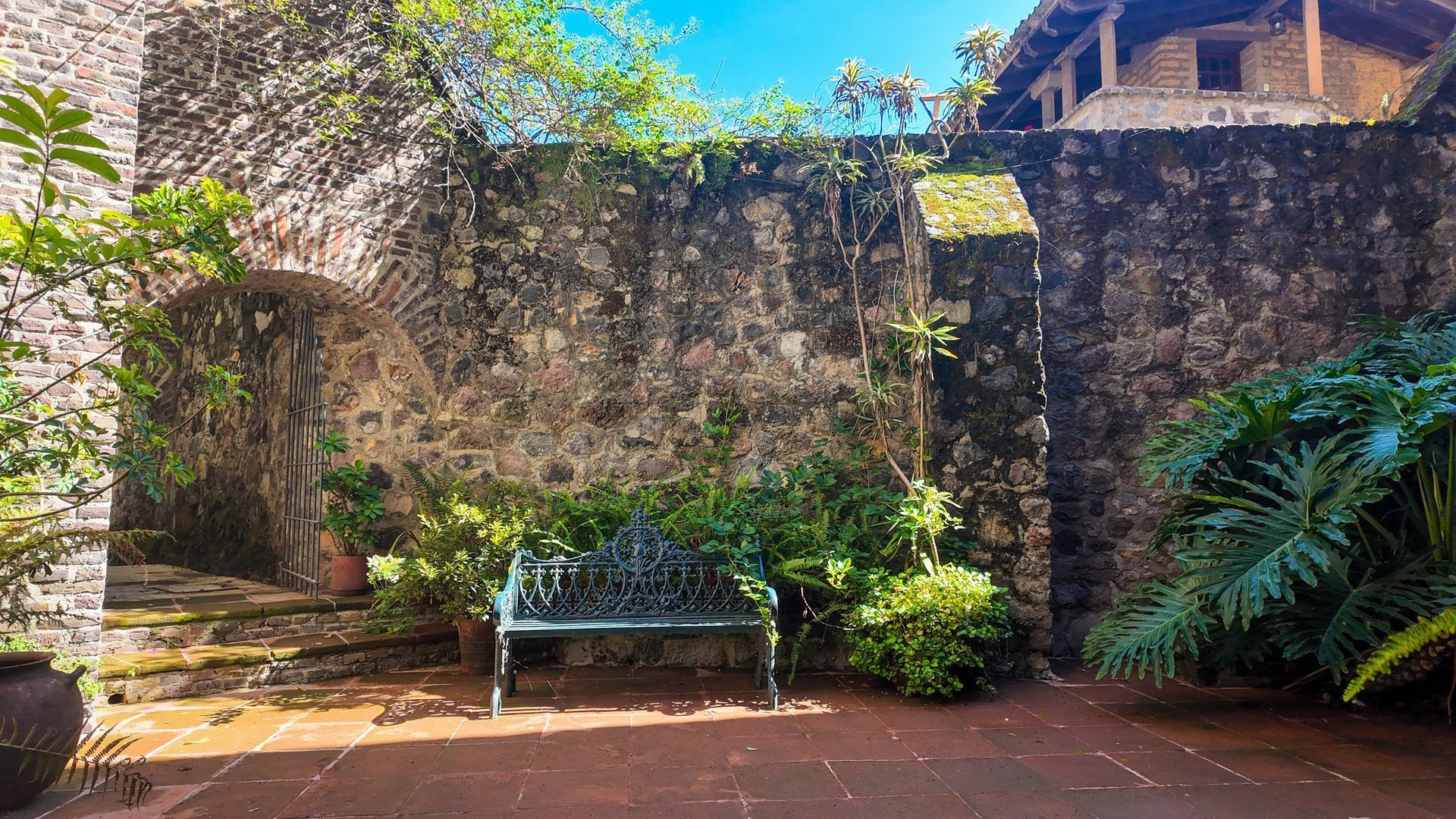 Patio con muro de piedra, banco verde, plantas en macetas y luz solar brillante bajo un cielo azul.