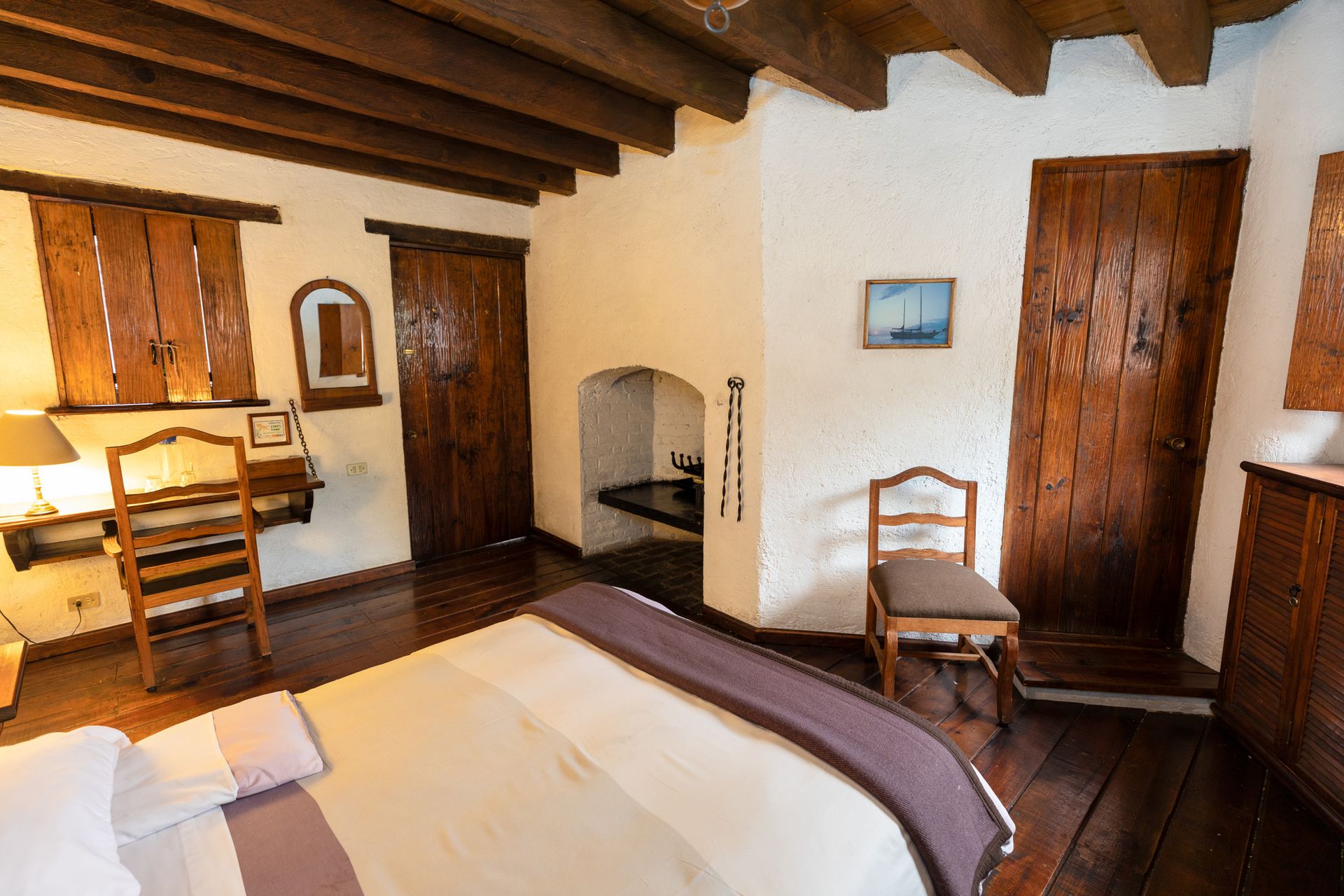 Dormitorio rústico con vigas de madera, cama, escritorio, chimenea y dos sillas.