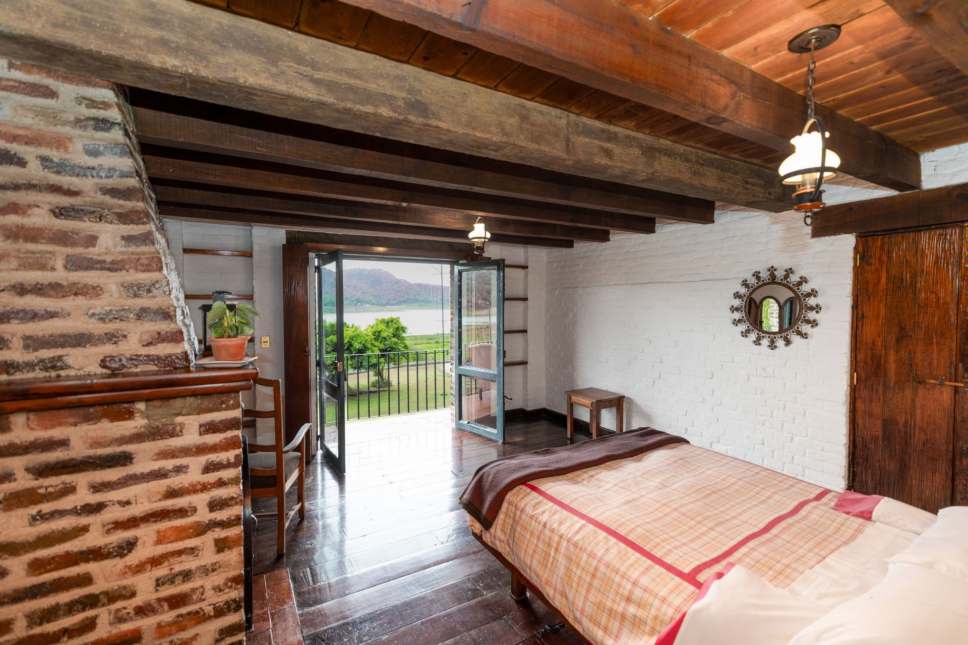 Habitación rústica con vigas a la vista, pared de ladrillo, cama y puertas francesas abiertas que dan a un balcón panorámico.