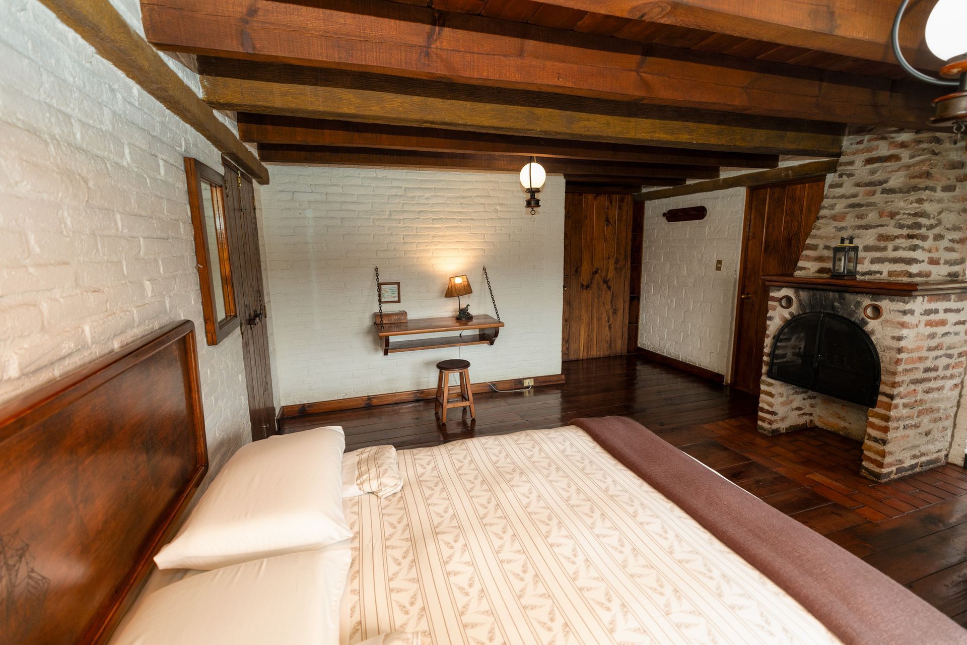 Dormitorio rústico con vigas de madera, chimenea de ladrillo y una cama con ropa de cama blanca.