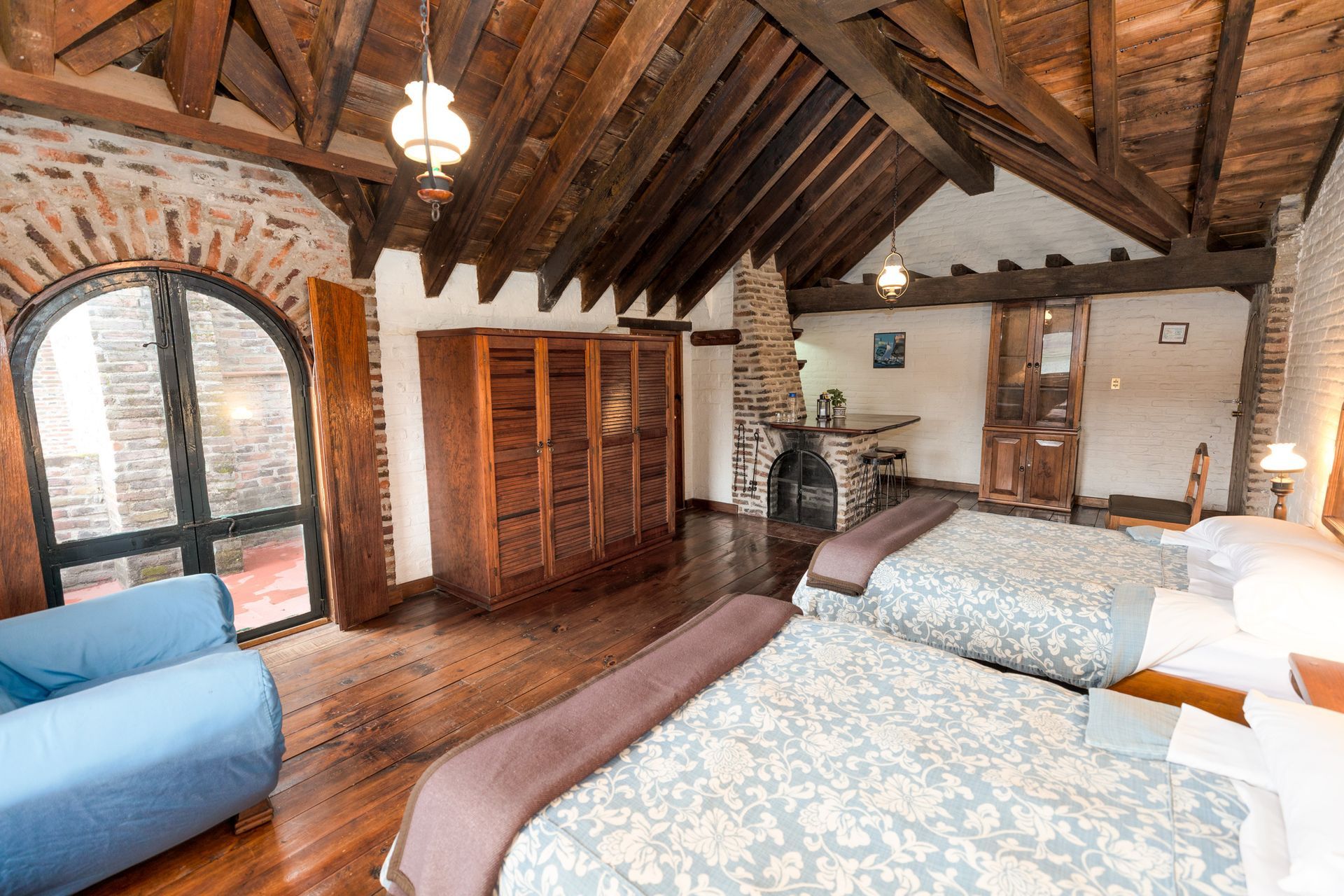 Habitación rústica con vigas de madera a la vista, ventanas arqueadas, dos camas individuales y chimenea.