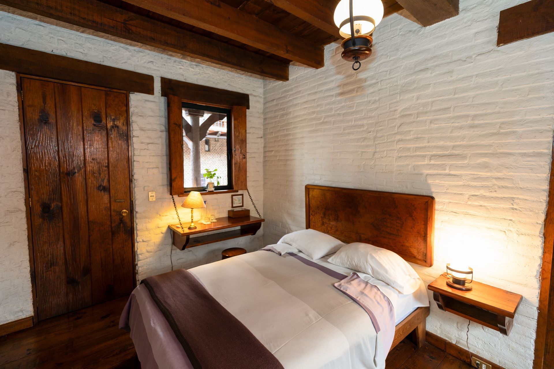 Dormitorio rústico con vigas de madera, paredes de ladrillo blanco y una cama bien hecha junto a lámparas de noche.