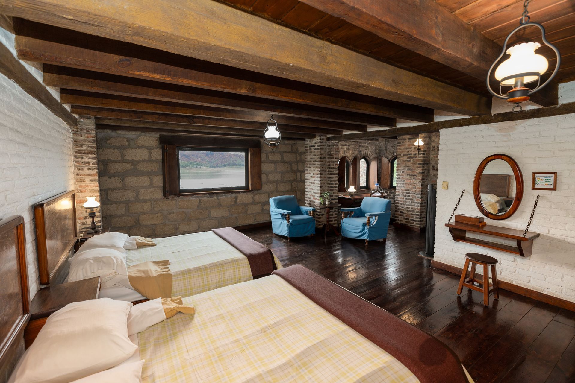 Habitación rústica con dos camas, paredes de piedra, vigas de madera y sillas azules cerca de una ventana.