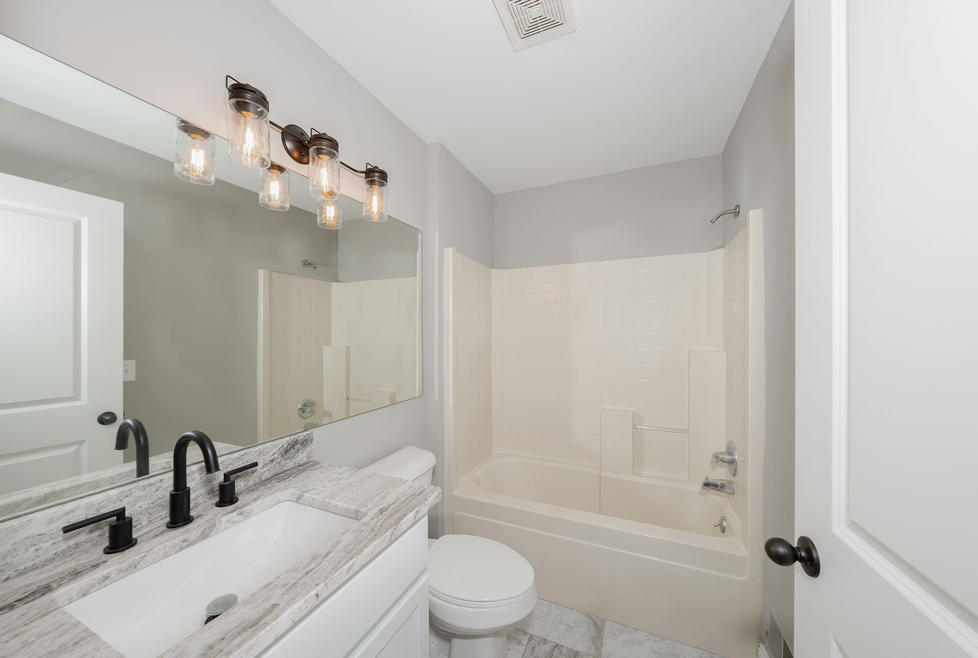 Bathroom with a white vanity, toilet, and bathtub. Features grey walls, a large mirror, and black fixtures.