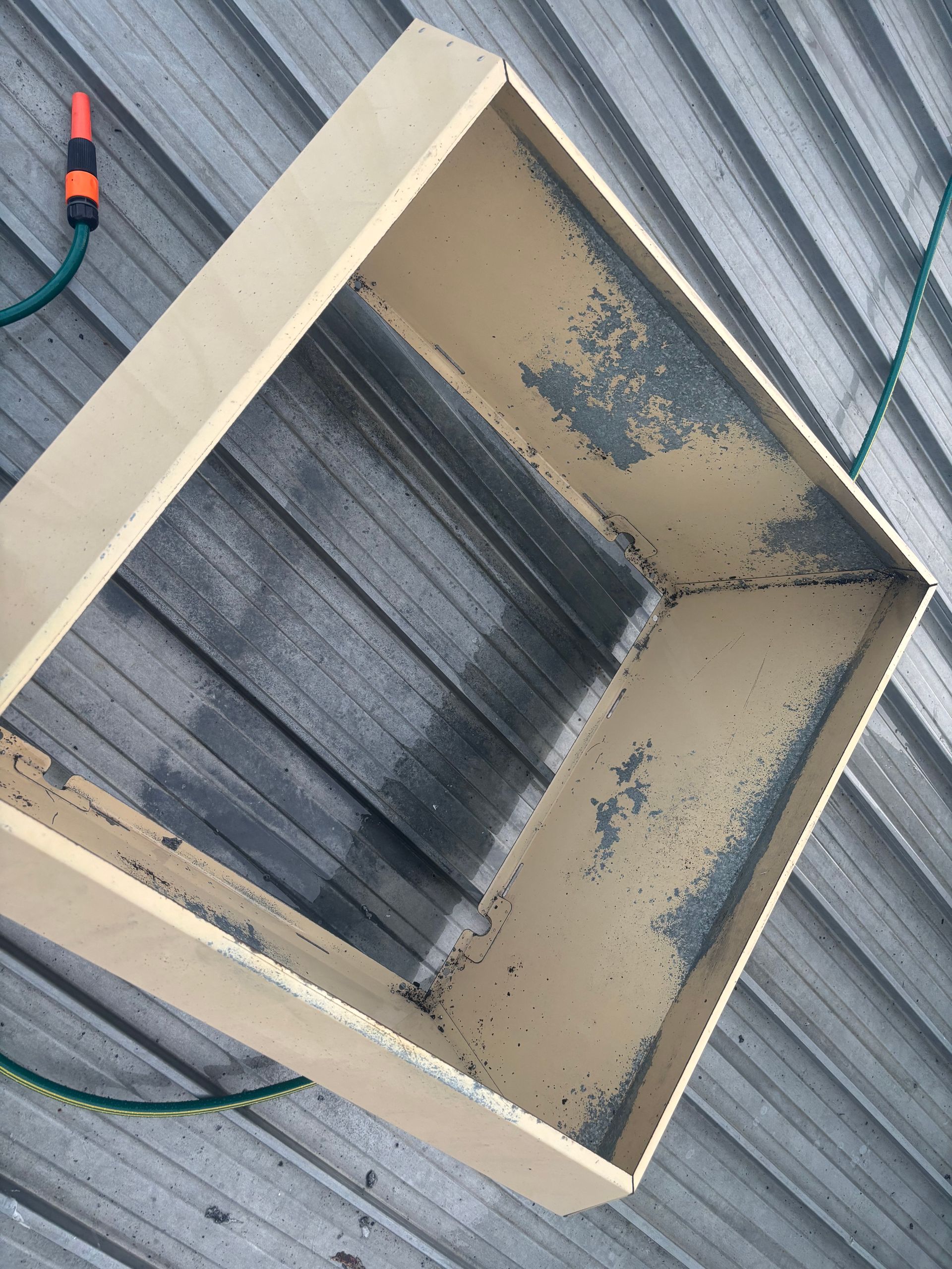 Square wooden box on a corrugated metal surface. Dark stains line the inside. A hose and nozzle are visible. - DM & MM Services, Exhaust Cleaning, Townsville, QLD.