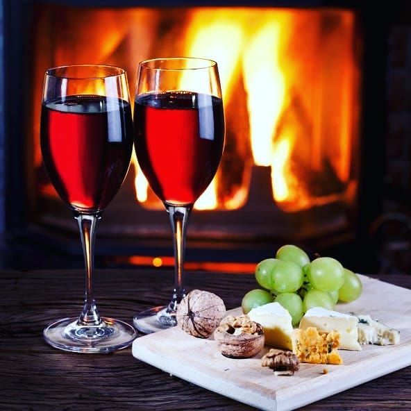 Two glasses of red wine are on a wooden table in front of a fireplace