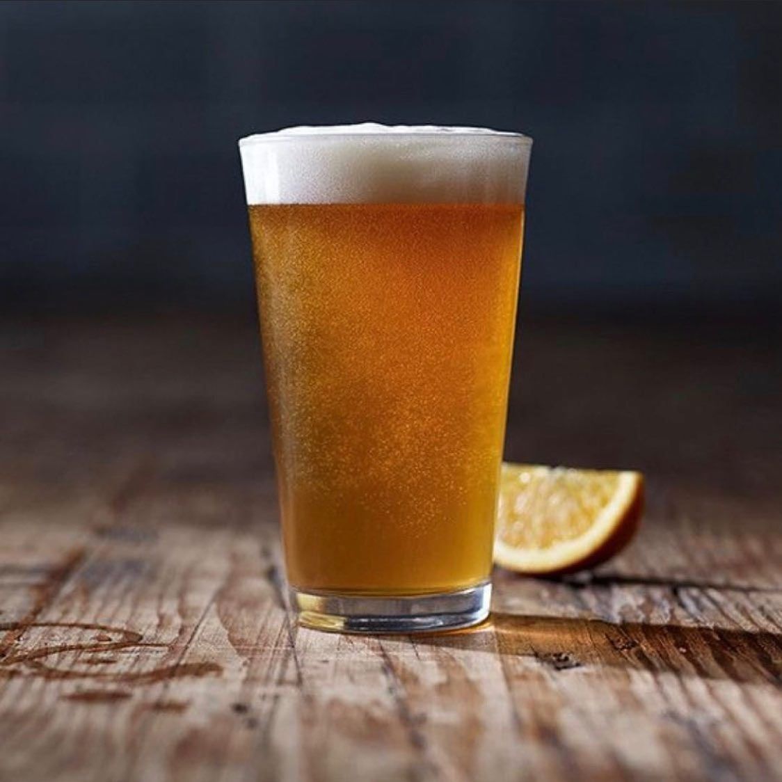A glass of beer is sitting on a wooden table next to an orange slice.