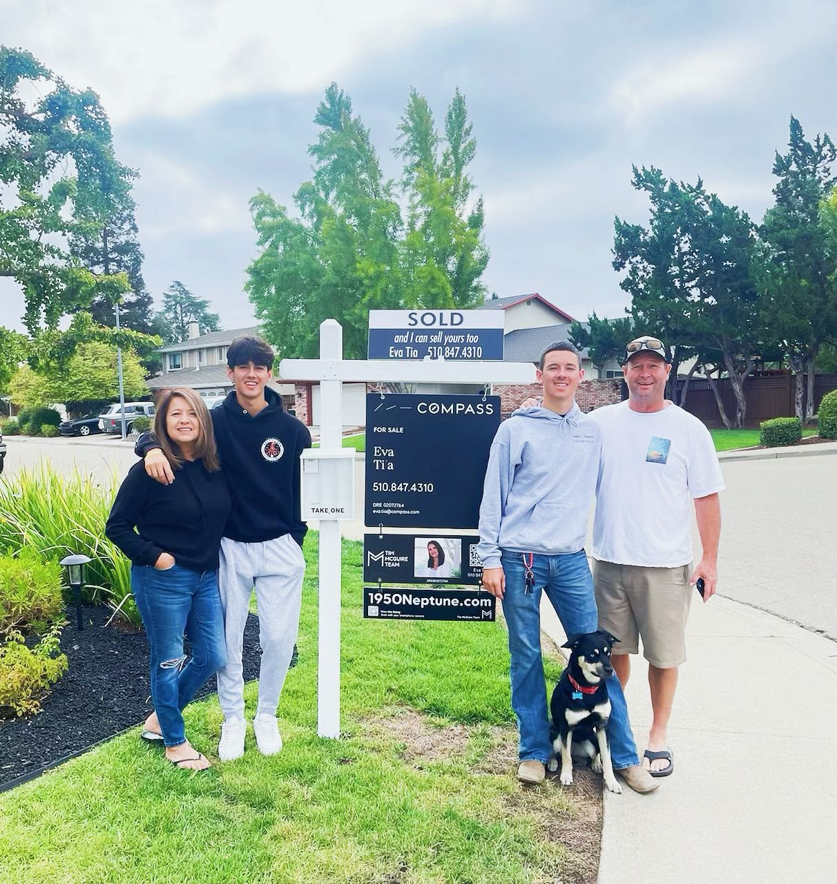 Four people and a dog pose beside a sold real estate sign on a suburban street.