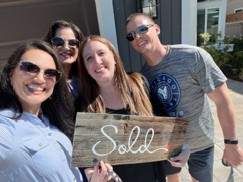 Four people smiling outside a house, holding a wooden “Sold” sign.