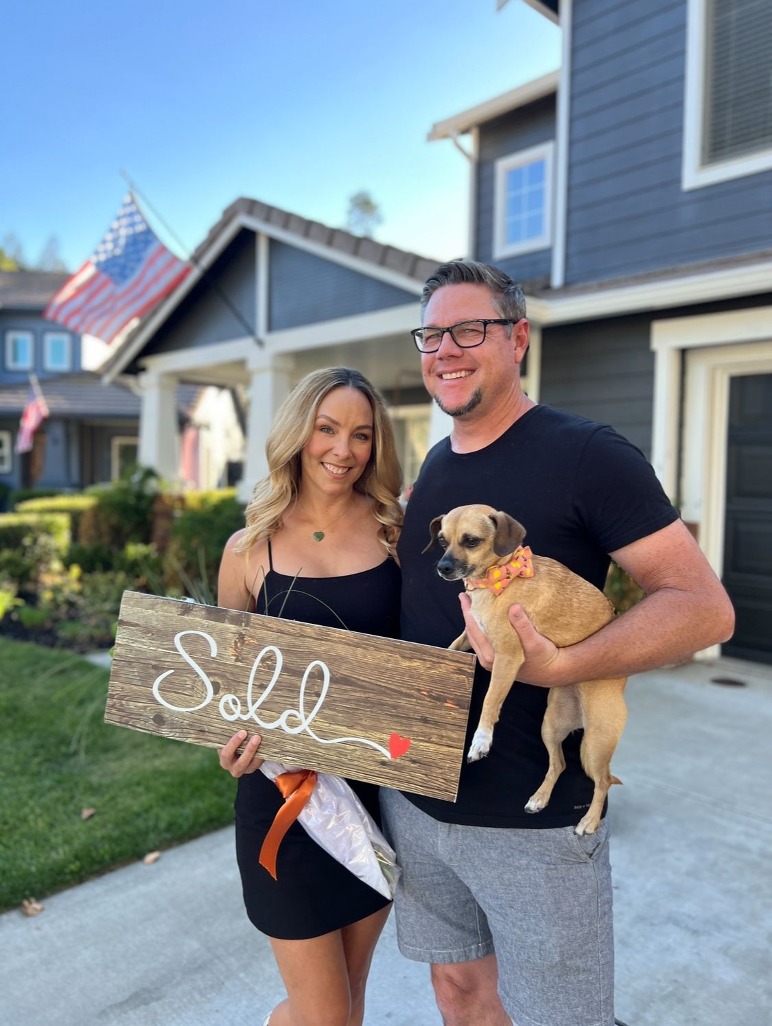Couple holding a “Sold” sign and a small dog in front of a house.