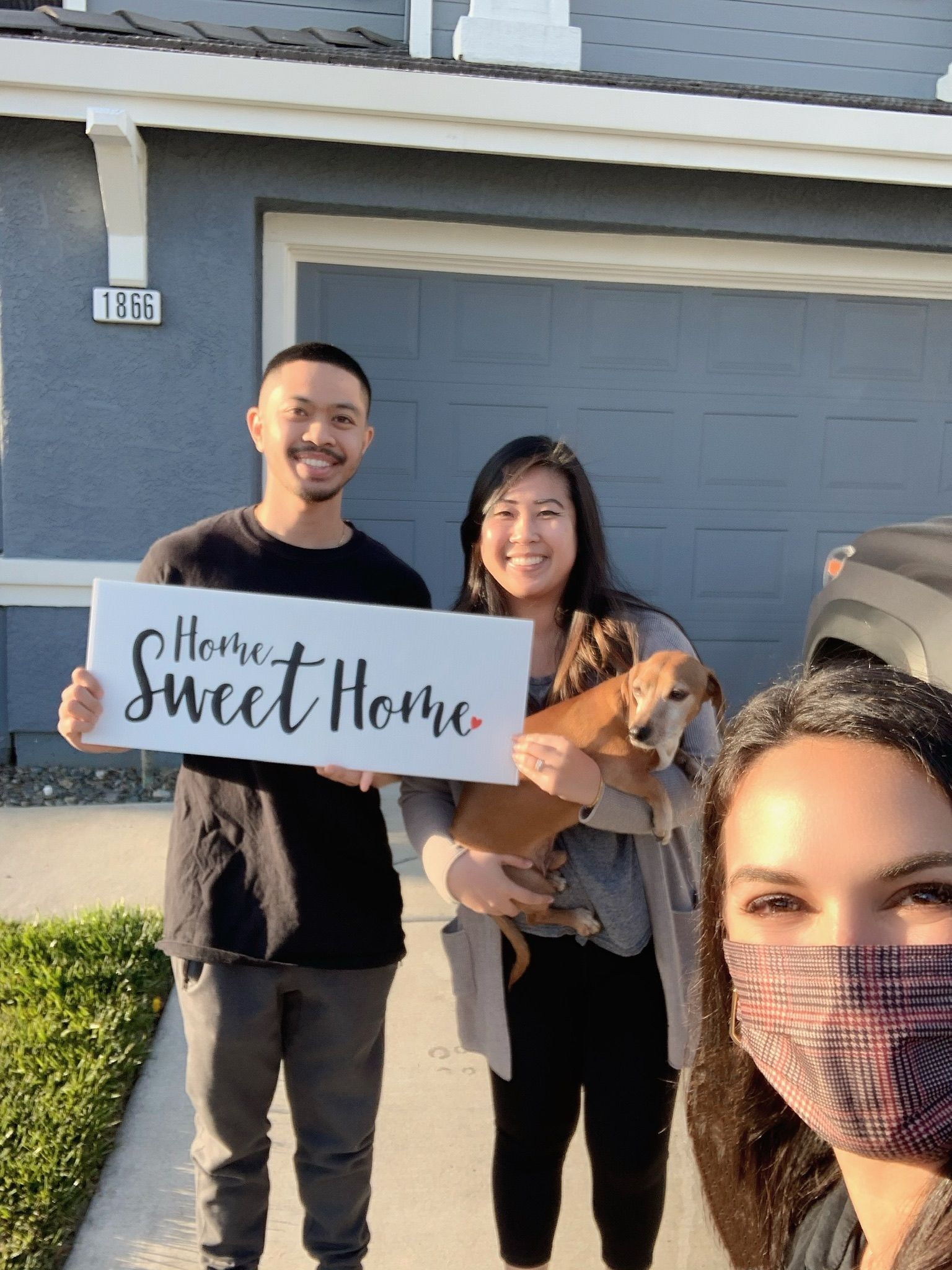 Three people posing outside a house, holding a “Home Sweet Home” sign and a dog.