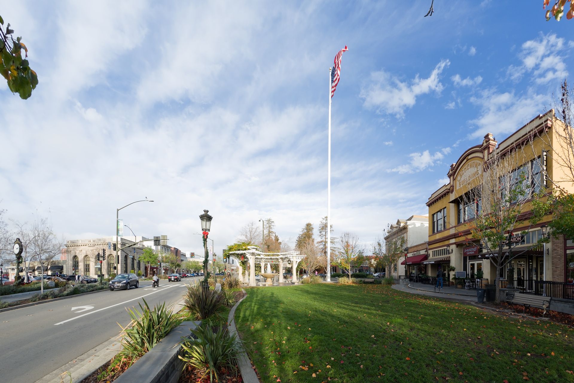 Wide street with a flagpole, landscaped median, and ornate buildings under a blue sky