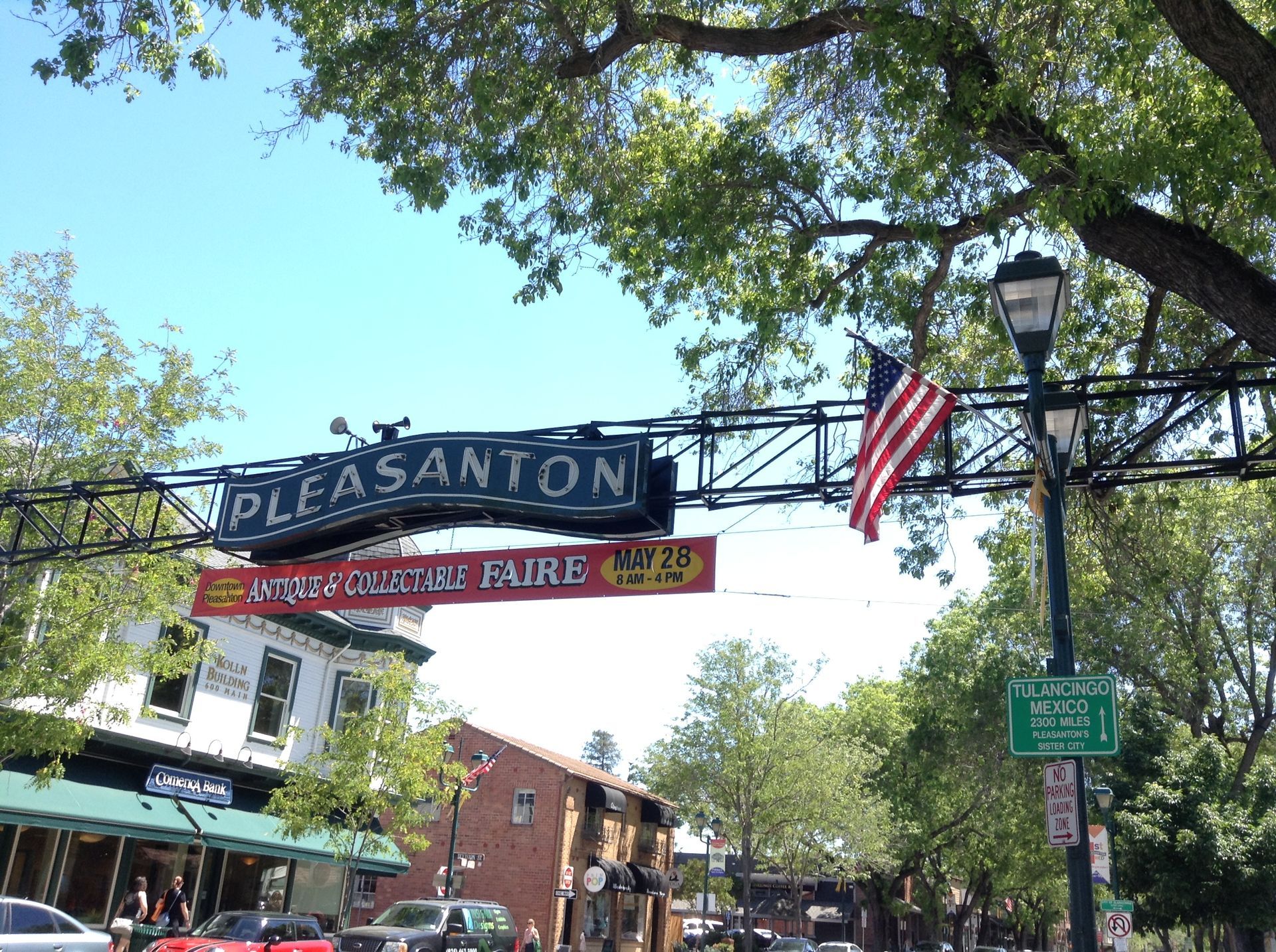 Downtown Pleasanton arch sign with hanging banners and an American flag under leafy trees