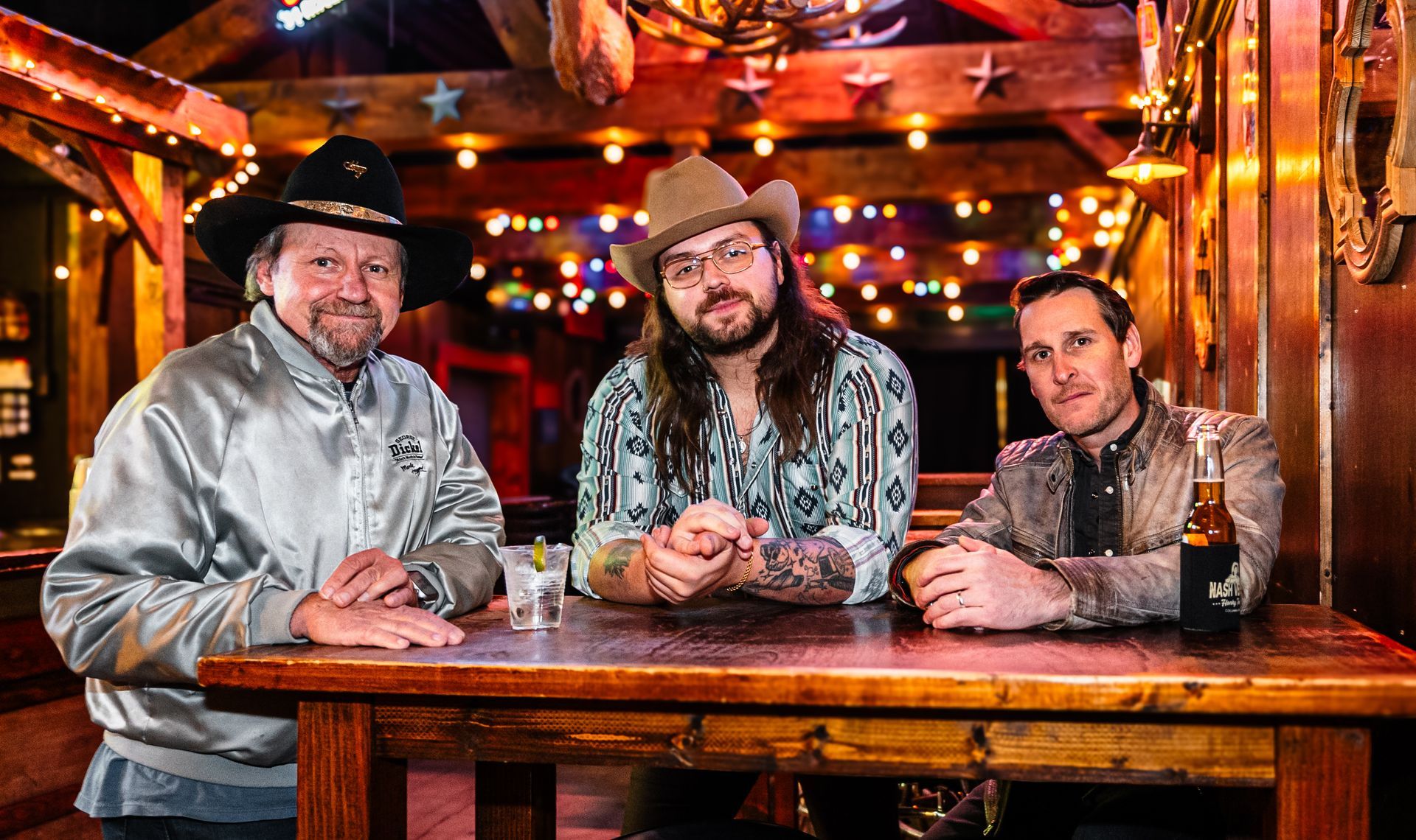 Three men in hats sit at a bar table in a warmly lit rustic pub with colorful lights.