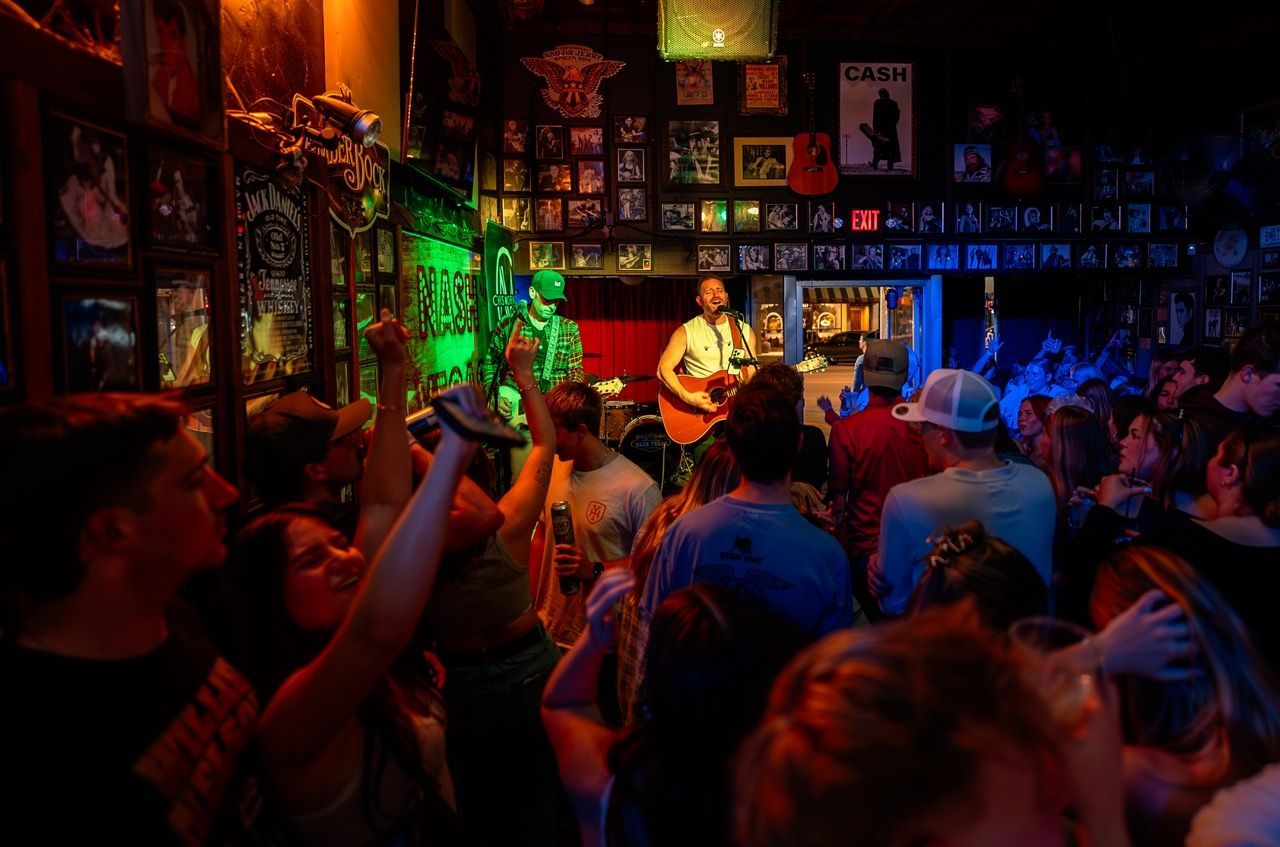 Crowded live music venue with colorful stage lights and audience raising hands toward the performers