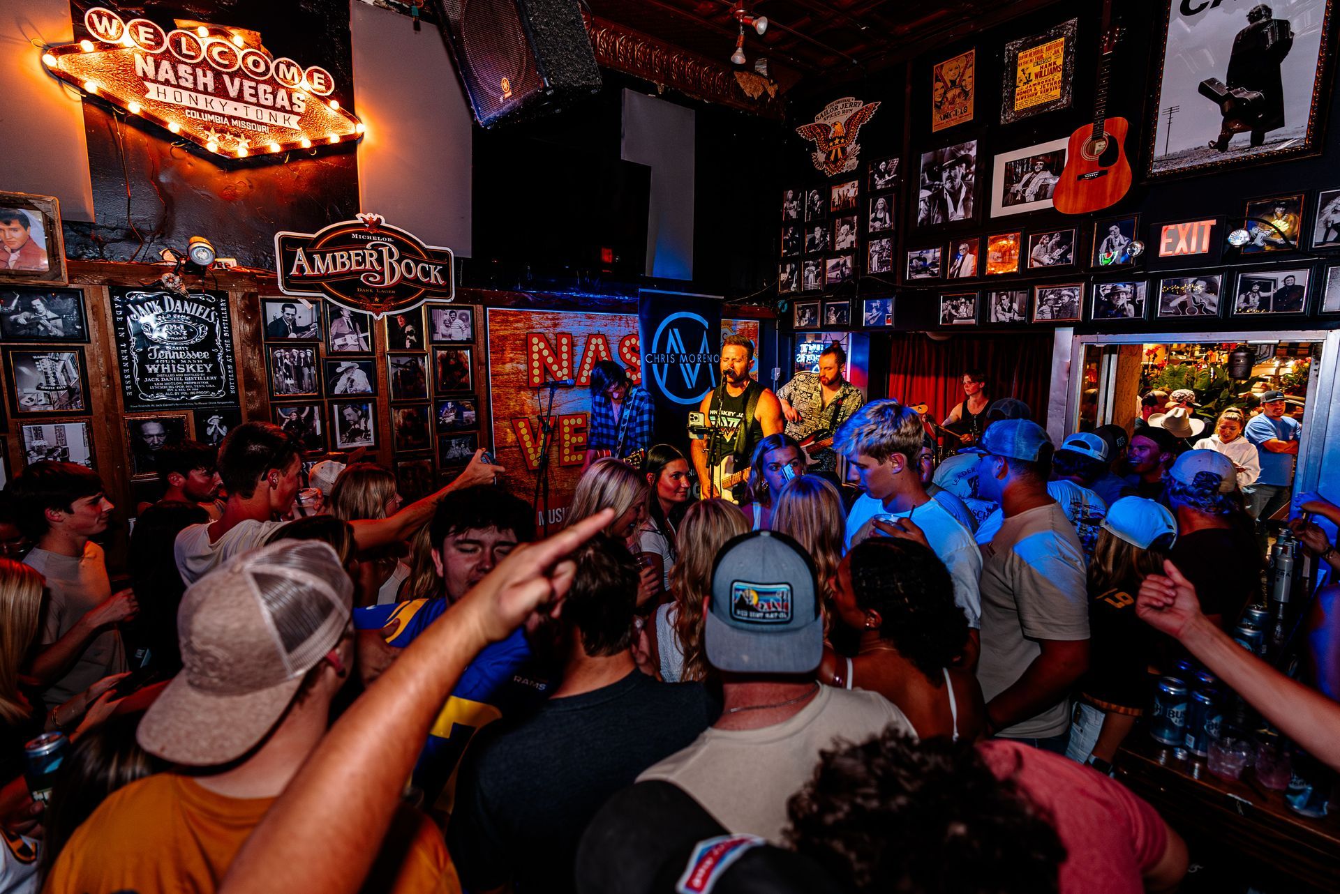 Crowded live music bar with a band onstage, colorful lights, and fans raising their arms