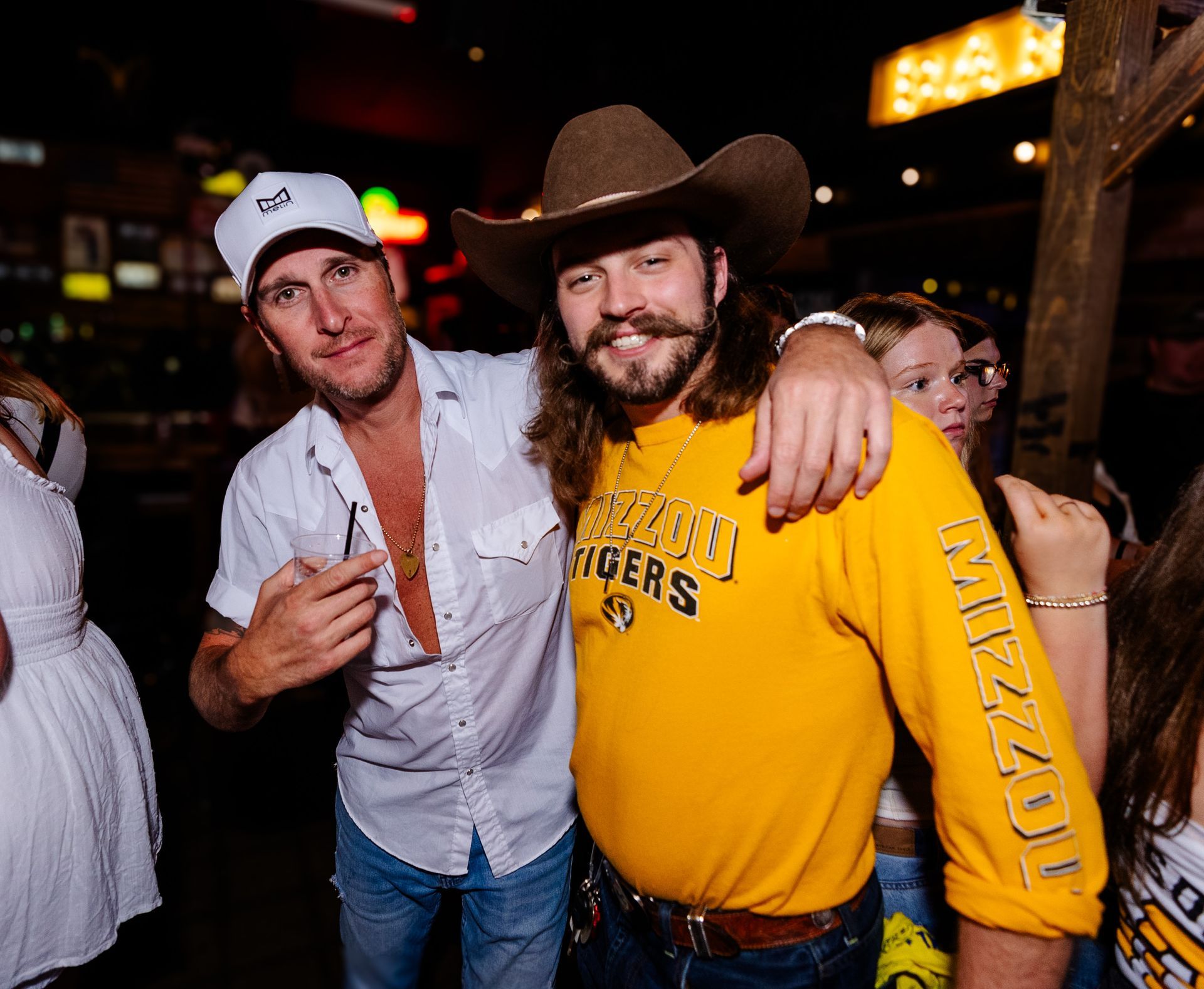 Two smiling men in cowboy hats at a lively bar, one in a yellow shirt and one in a white shirt.