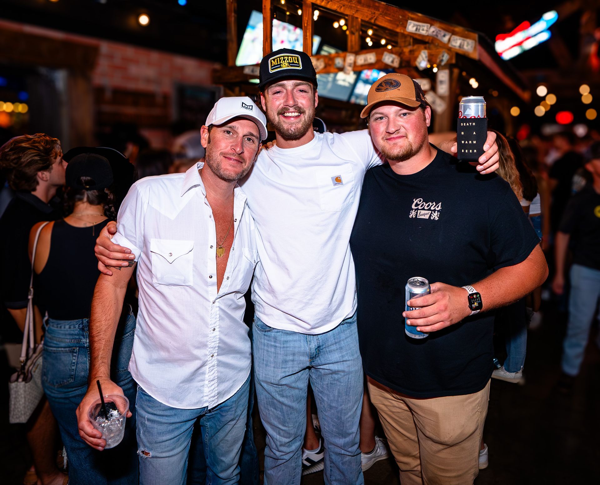 Three men smiling and posing together at a nighttime bar with drinks in hand