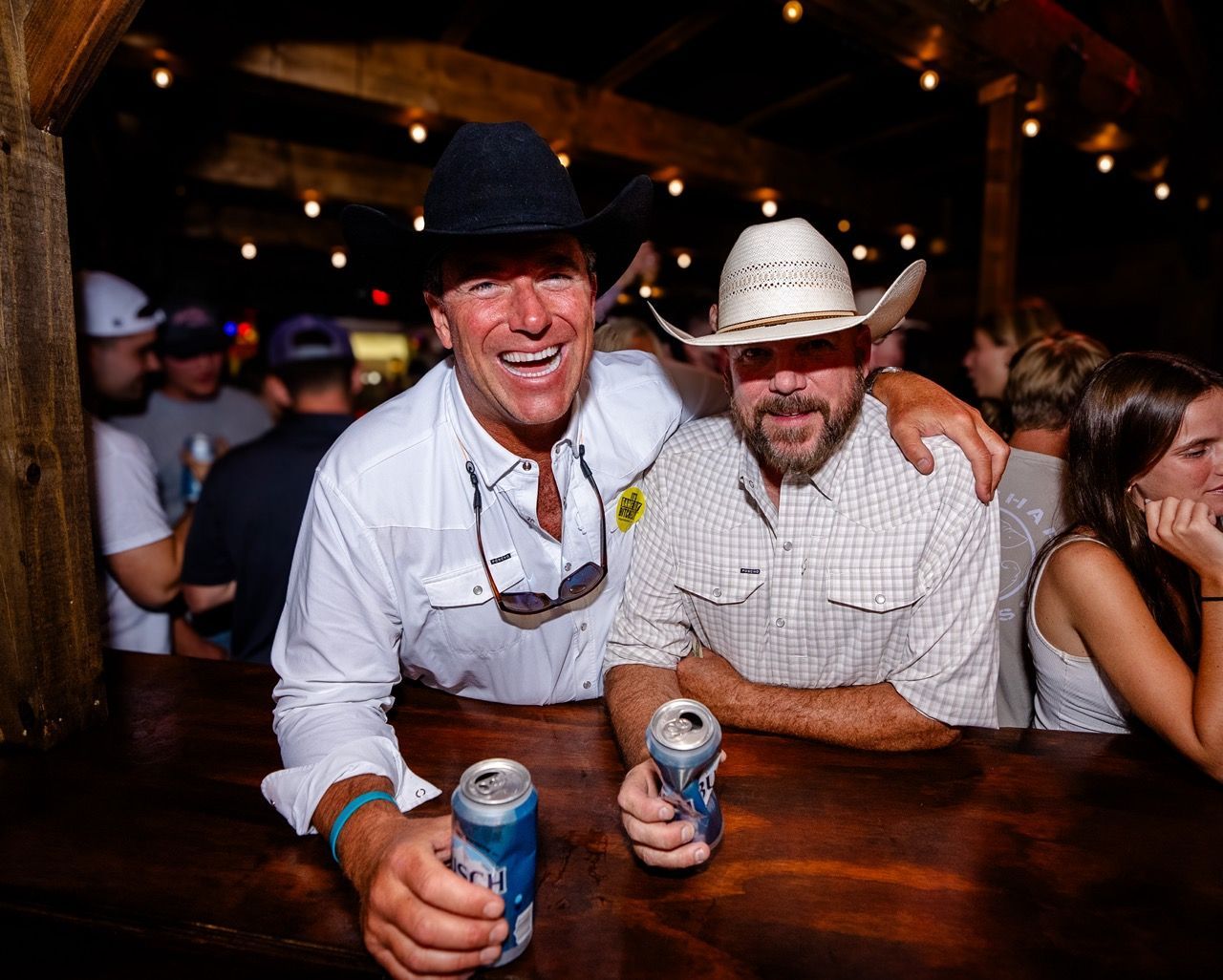 Two smiling men in hats holding drinks at a dimly lit bar, leaning on a wooden table