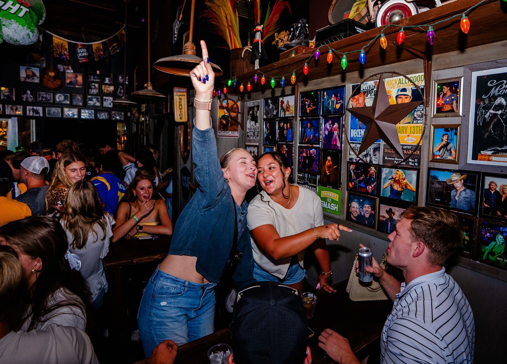 People dancing and celebrating in a colorful, crowded bar with neon lights and wall photos