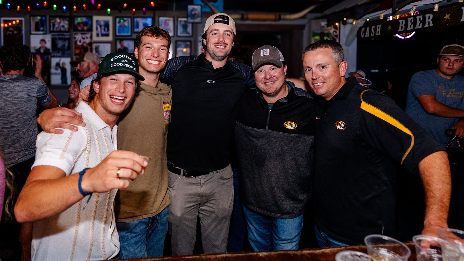 Five people smiling and posing together at a bar, holding drinks under colorful string lights.