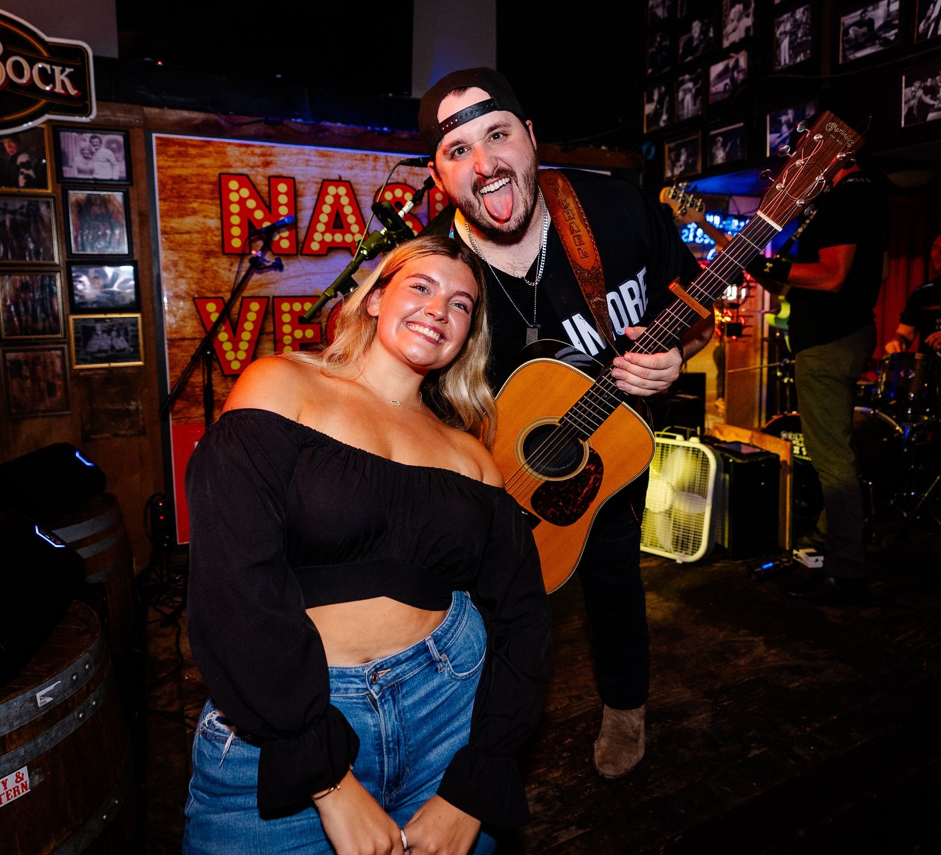 Two smiling people posing with a guitar in a dimly lit bar with rock decor and neon signs