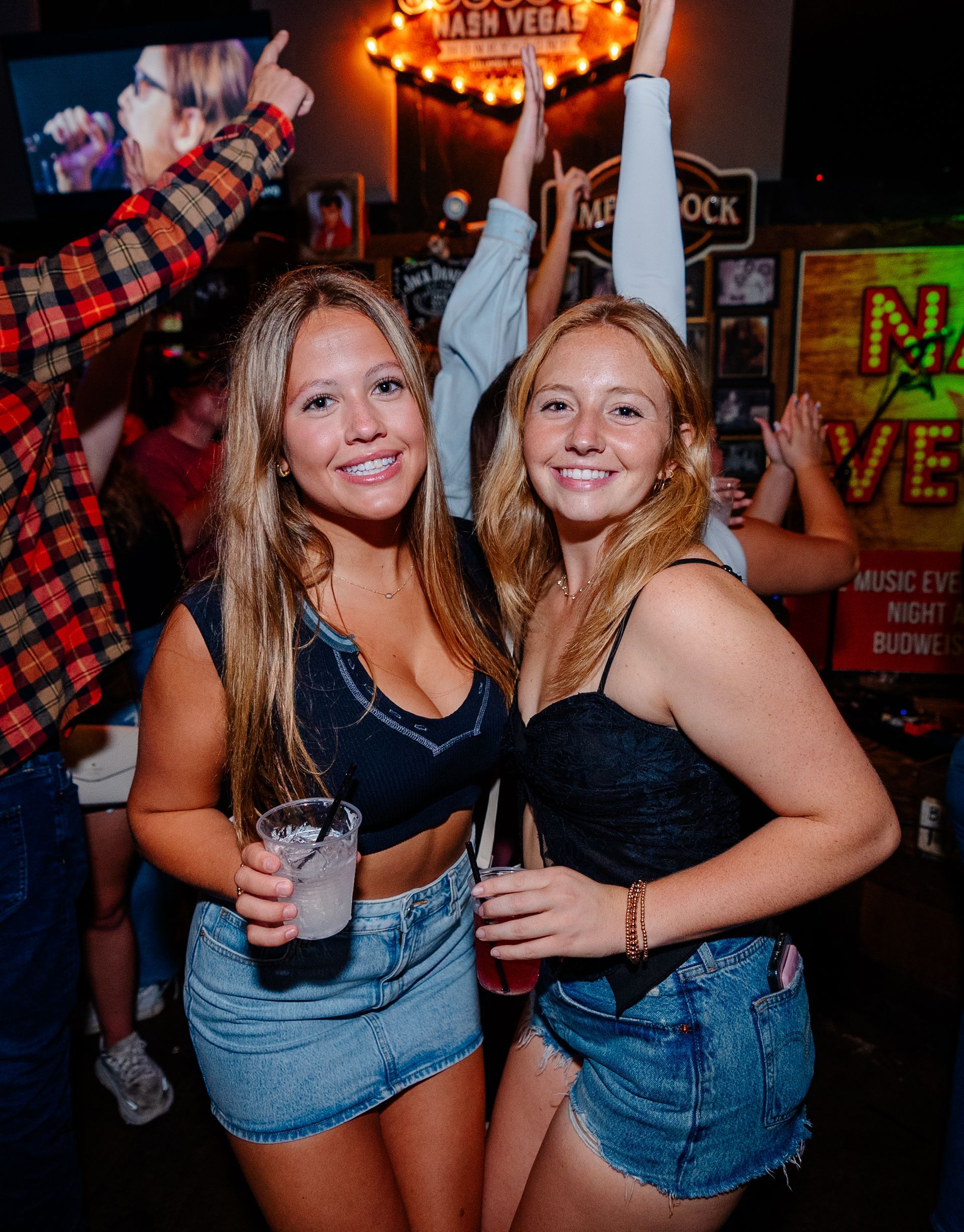 Two women dancing in a crowded bar, smiling and holding drinks, with colorful neon signs in the background.