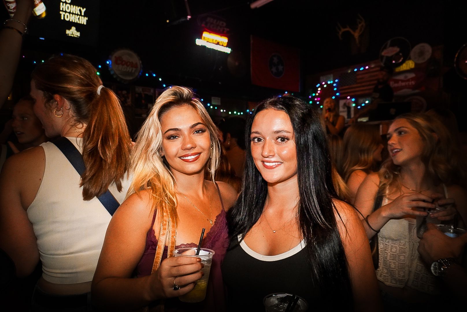 Two women smiling at a dimly lit bar, one holding a drink, with colorful lights in the background