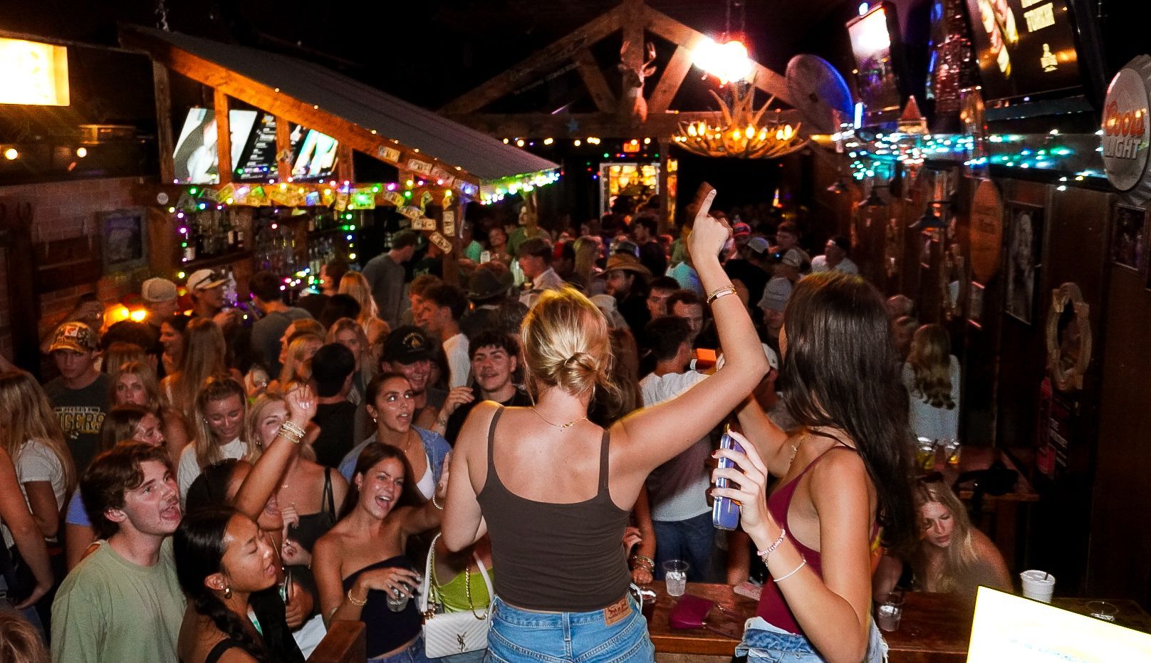 Crowded nightclub scene with people dancing, raised drinks, and colorful lights under wooden beams