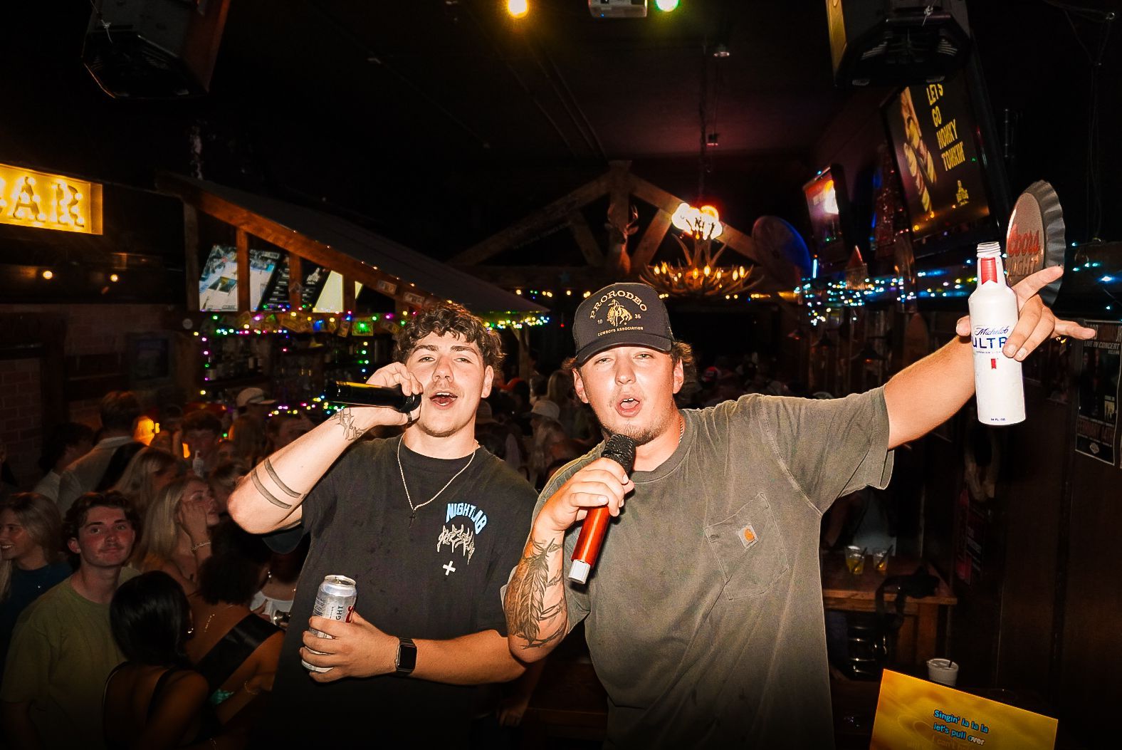 Two people singing into microphones in a dim bar, one holding a drink, crowd in the background.
