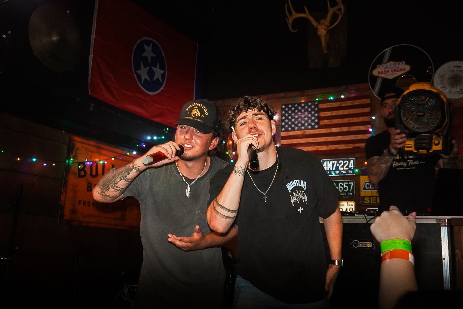 Two performers singing onstage in a bar, with a crowd and Tennessee flag in the background