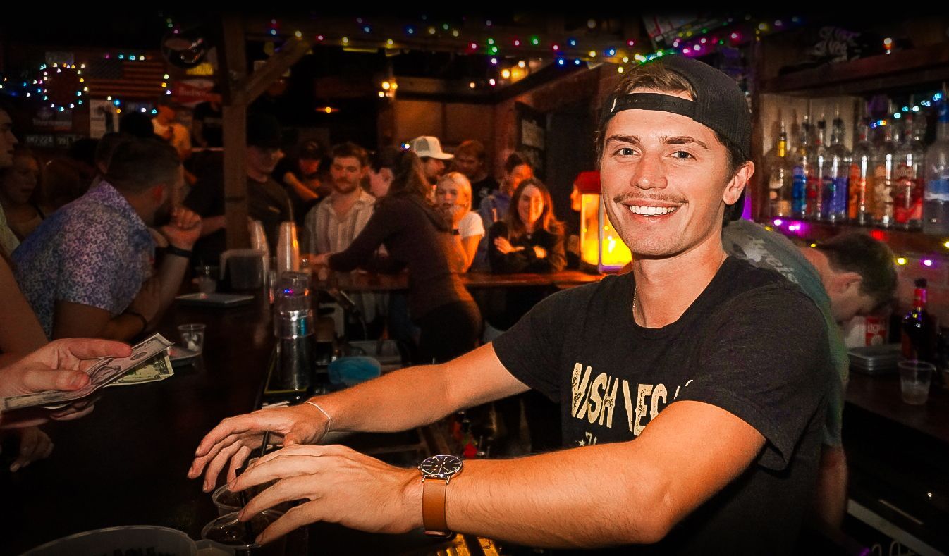 Smiling bartender in a black shirt serving drinks in a lively bar with colorful lights